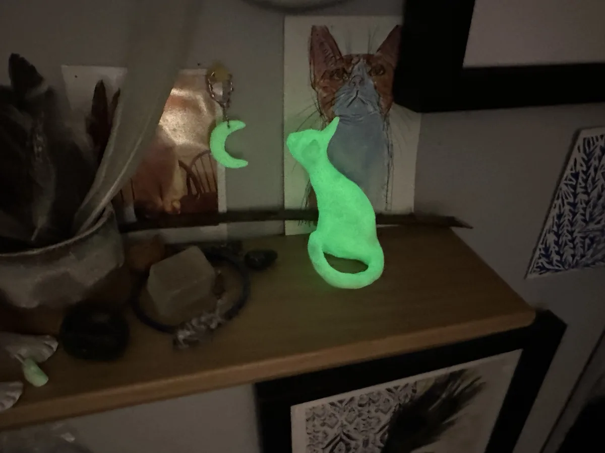A glowing green cat-shaped figurine sits on a wooden shelf in a dimly lit room, facing a painting of a cat. Beside it, a…