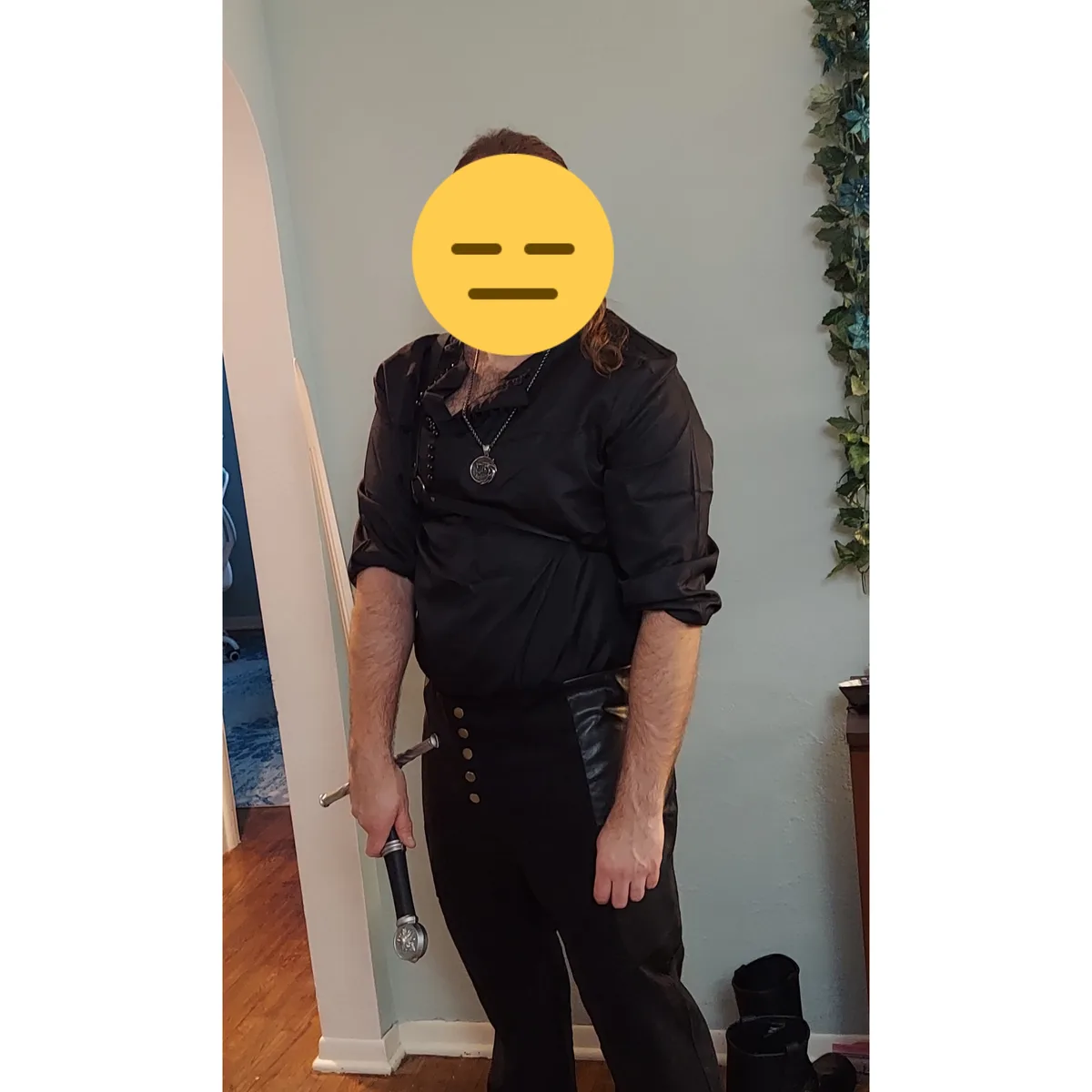 A person in a dark, fantasy-themed outfit with a sword, wearing a neutral face emoji over their face, stands indoors. They…