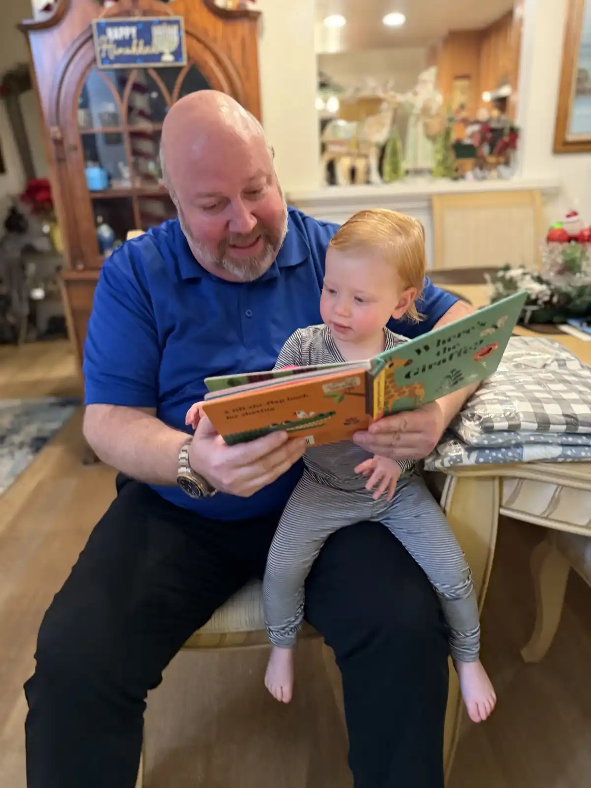 A bald man with a beard in a blue shirt sits on a chair holding a young child on his lap while reading a children's book…