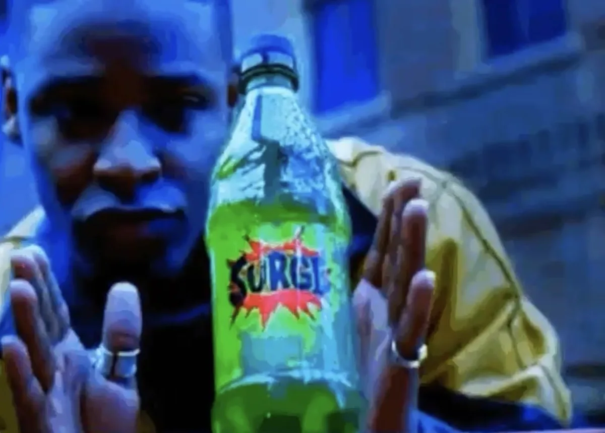 A man in a yellow jacket holds a green bottle of Surge soda, looking intensely at the camera with both hands outstretched…
