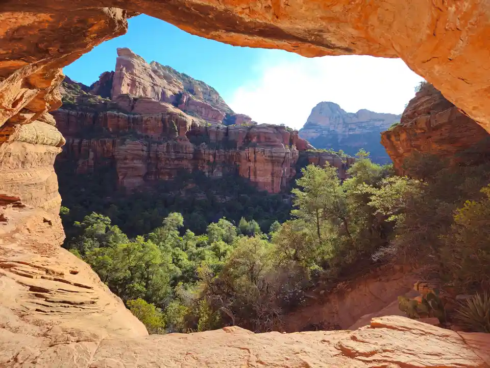 A scenic view from inside a natural sandstone arch, looking out onto red rock canyon walls and lush green trees under a…
