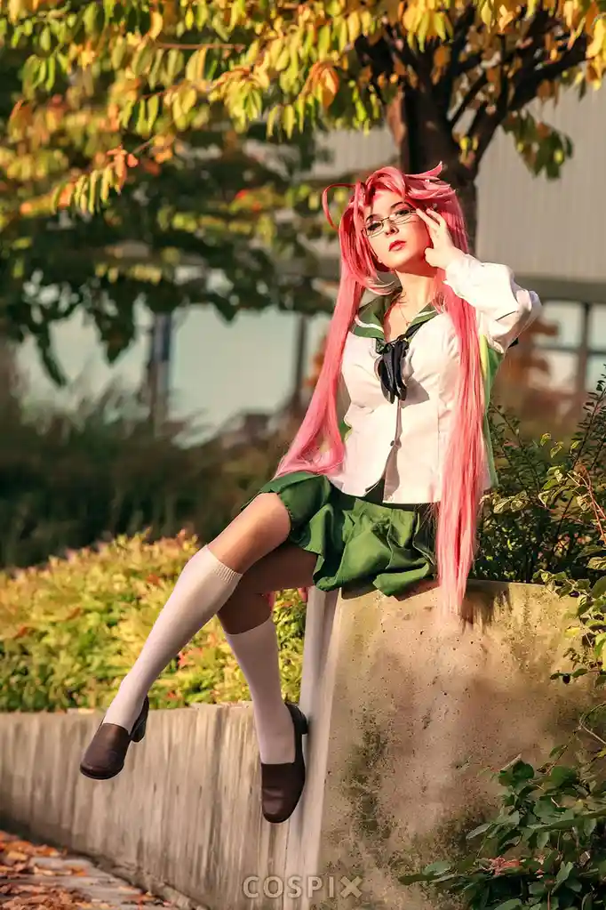 A person with long pink hair and glasses is cosplaying as a character in a green and white school uniform, sitting on a…