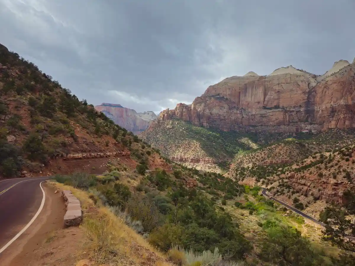 A scenic view of a winding road through a canyon with red rock cliffs and green vegetation under a cloudy sky. The overlaid…