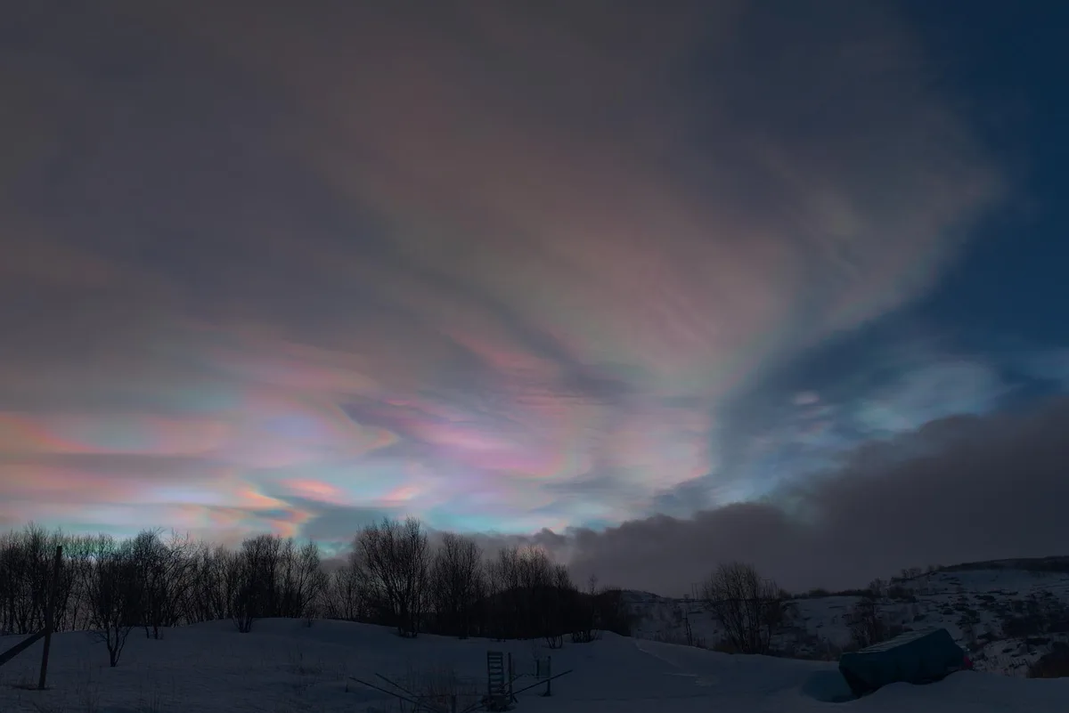 A snowy landscape at dusk or dawn with silhouetted trees and a snow-covered hill, under a sky displaying iridescent…