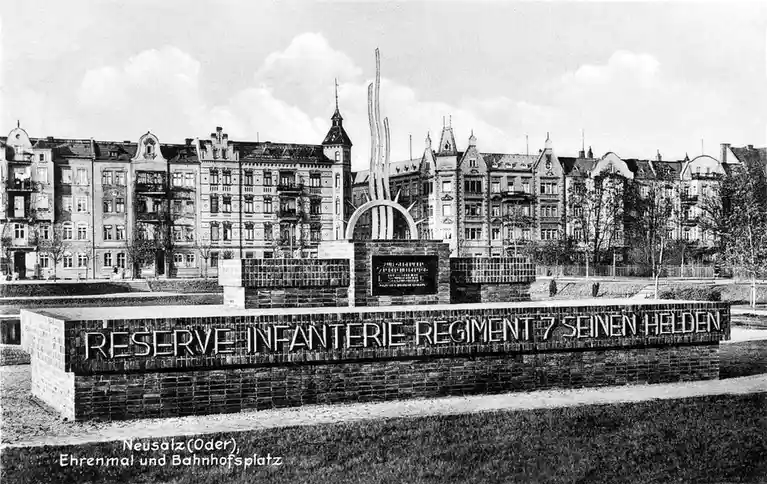A black-and-white historical photograph shows a memorial monument in a town square, with ornate buildings in the background…