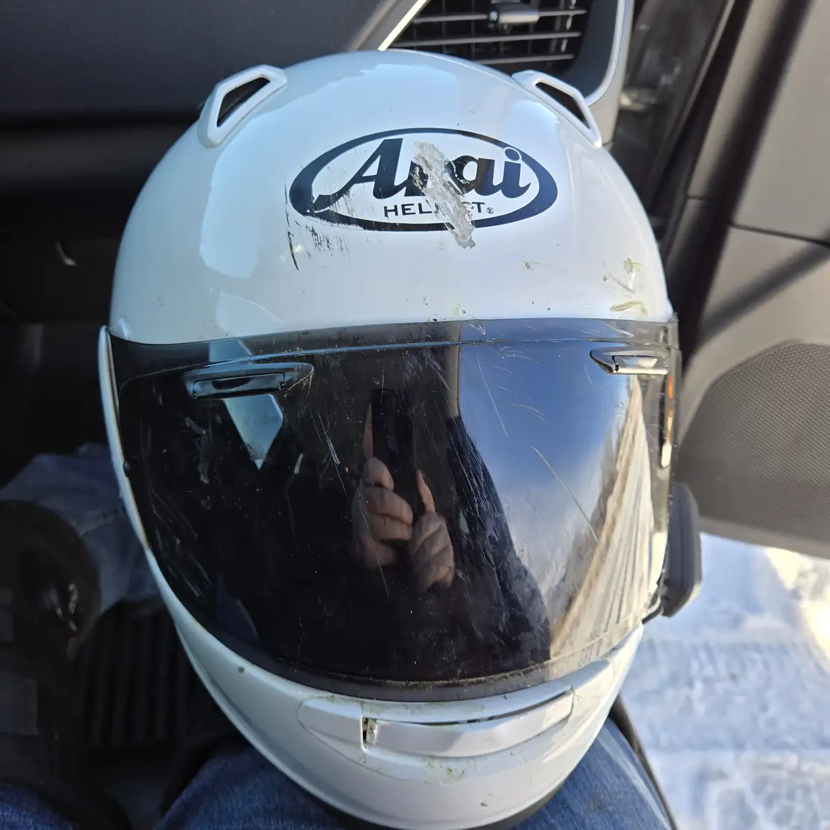A white Arai motorcycle helmet sits inside a car, visibly scratched and dirty with a large chip in the logo. The visor is…