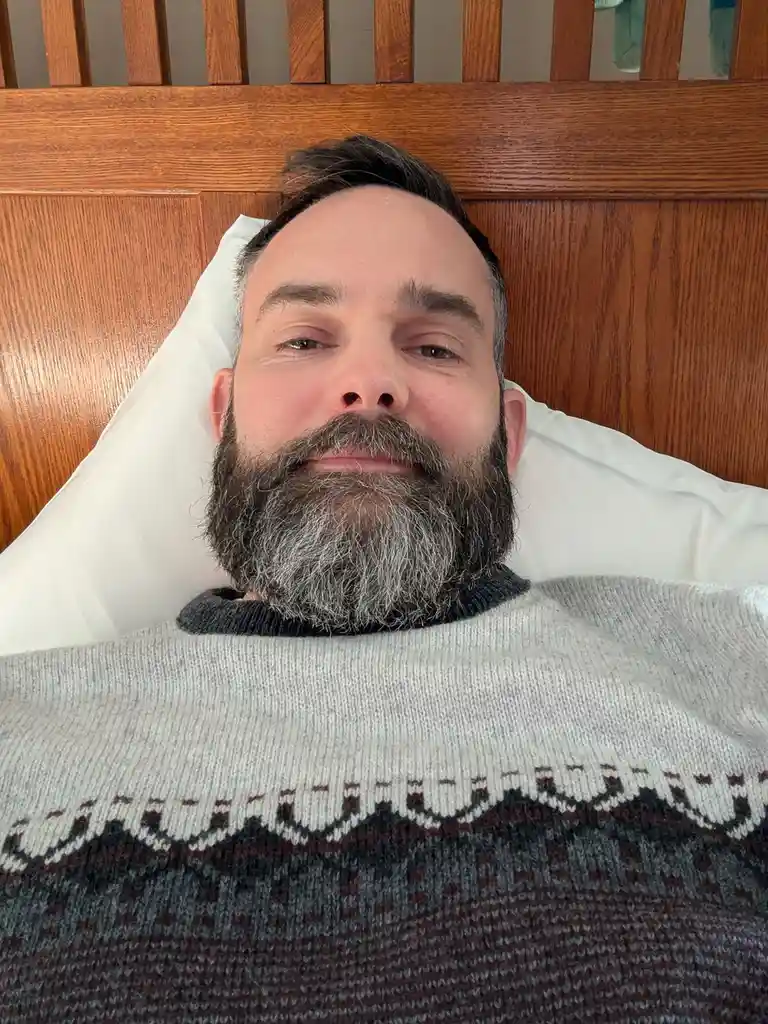 A man with a full, graying beard lies in bed, propped up on a white pillow against a wooden headboard, wearing a gray and…