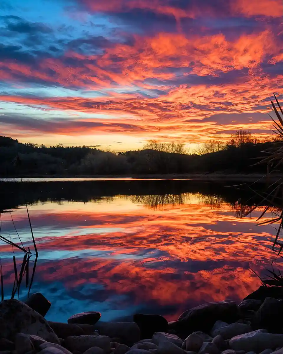 A serene landscape photo of a lake at sunset, with vibrant orange and blue clouds reflected in the water, framed by rocks…
