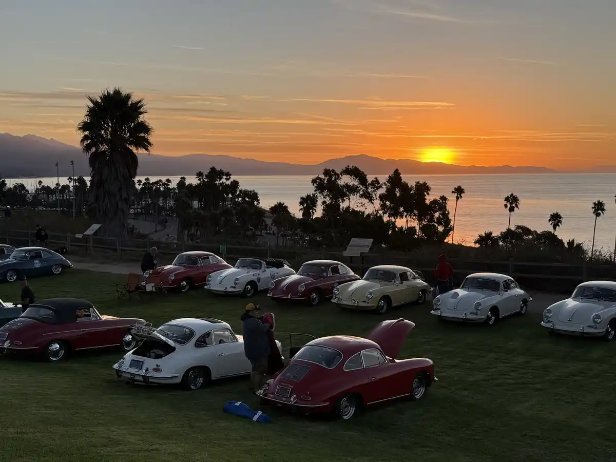A scenic photo of multiple vintage Porsche 356 cars parked on grass overlooking a sunset over water with palm trees and…