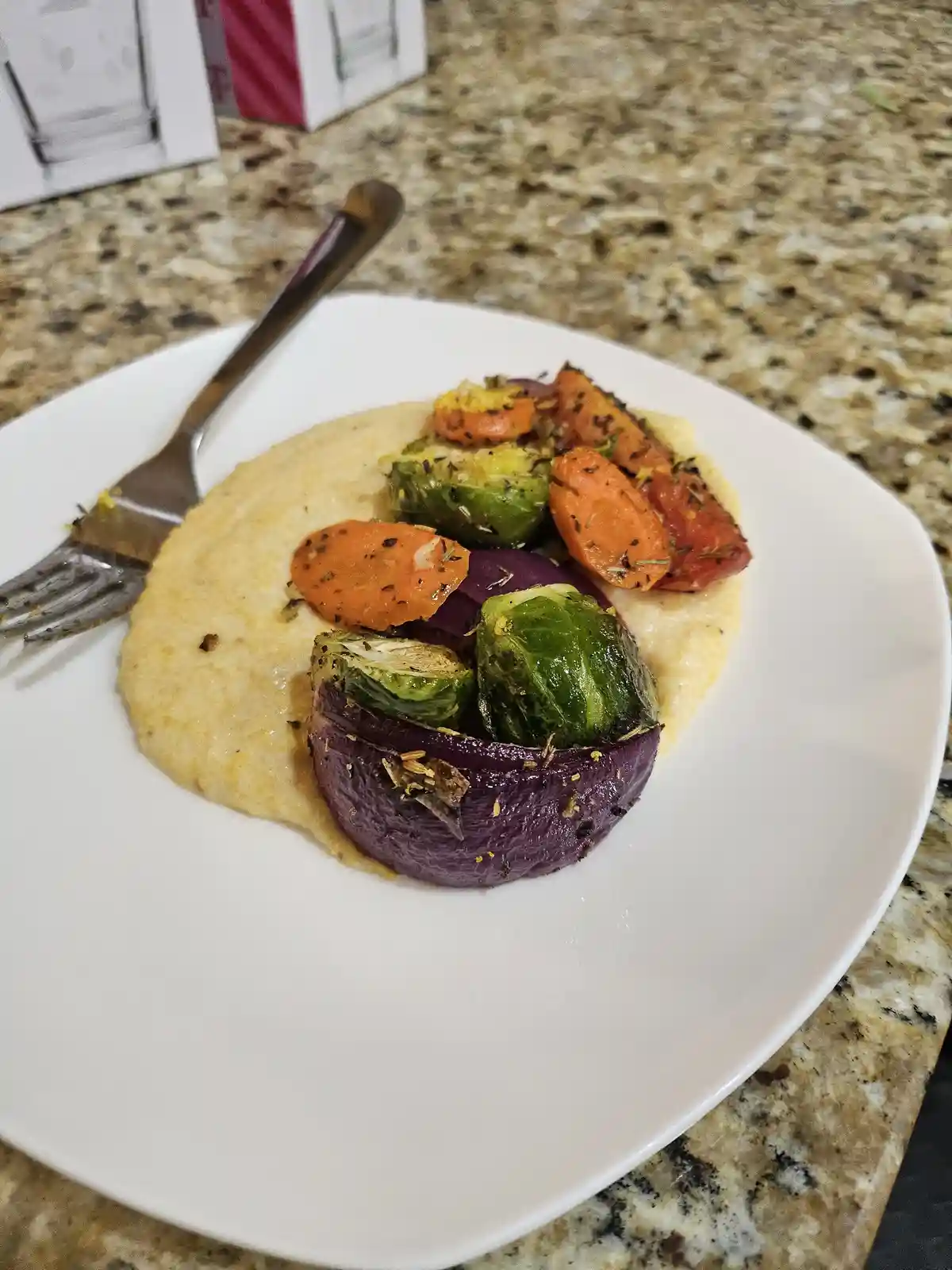 A white plate holds a bed of creamy polenta topped with roasted vegetables including orange carrots, green brussels sprouts…