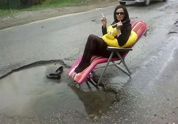 a woman wearing sunglasses and black tights sits in a pink and red striped folding chair placed in a large pothole filled…