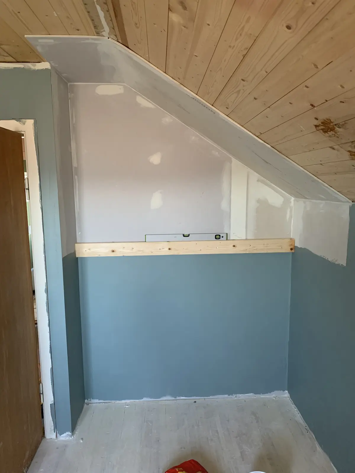A room under renovation with a sloped ceiling and partially painted walls, featuring a wooden plank with a level tool…