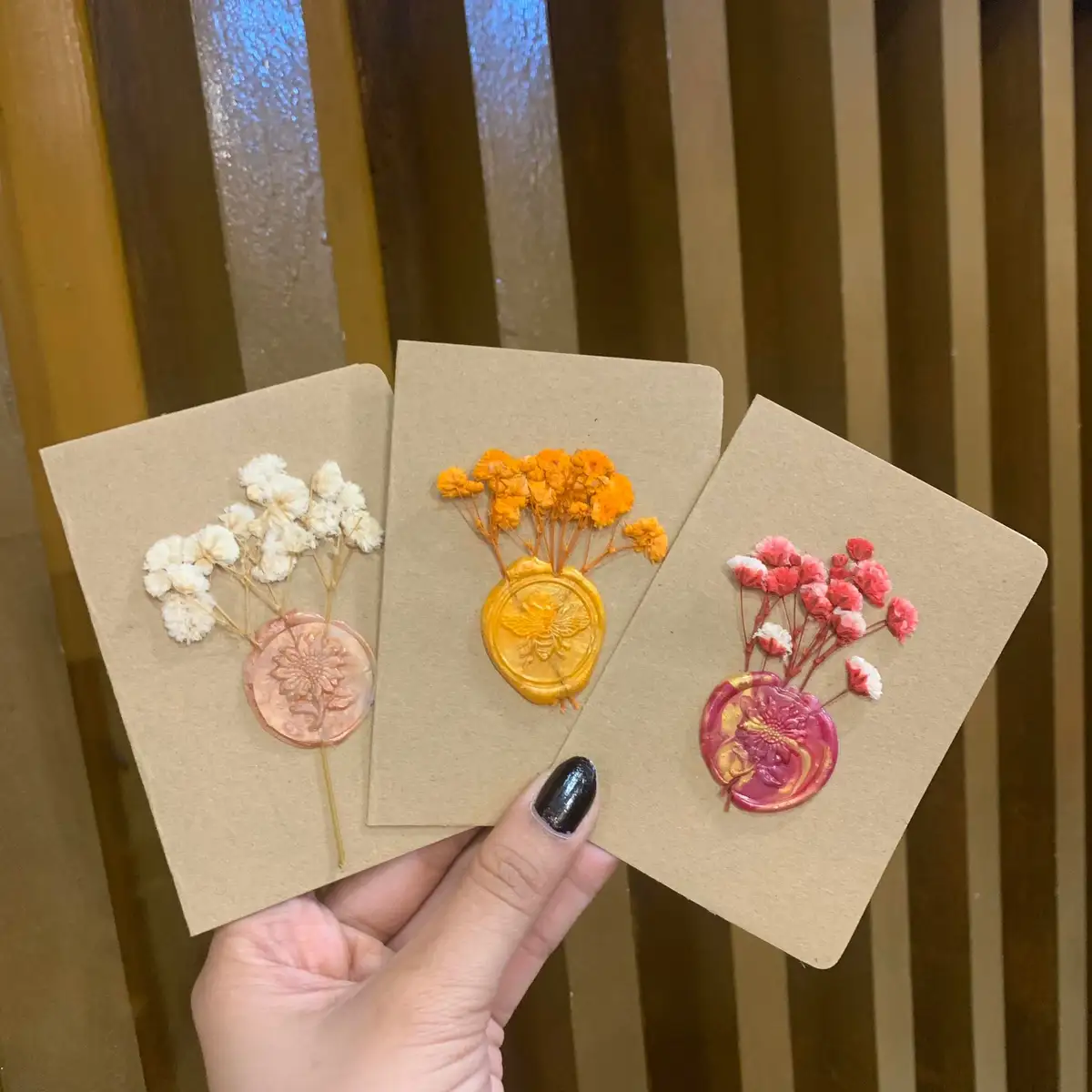 A hand holds three brown paper cards, each adorned with dried flower sprigs and a colorful wax seal. The left card has white…