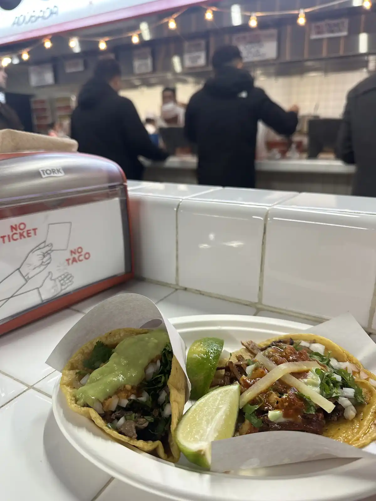 A close-up photo shows two tacos on a white paper plate with lime wedges, in front of a food stall counter. To the left, a…