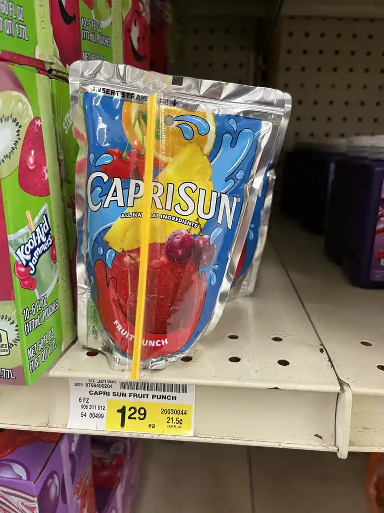 A Capri Sun fruit punch pouch sits on a store shelf with a yellow straw inserted, priced at $1.29. The packaging features…