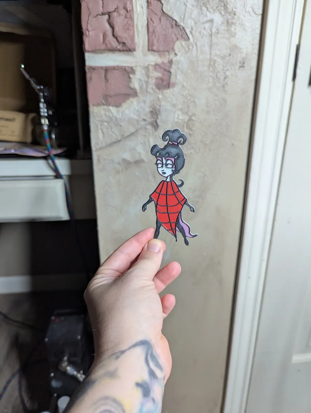 A hand holds up a cutout of a cartoonish female character with black hair in a bun, glasses, and a red spiderweb-patterned…
