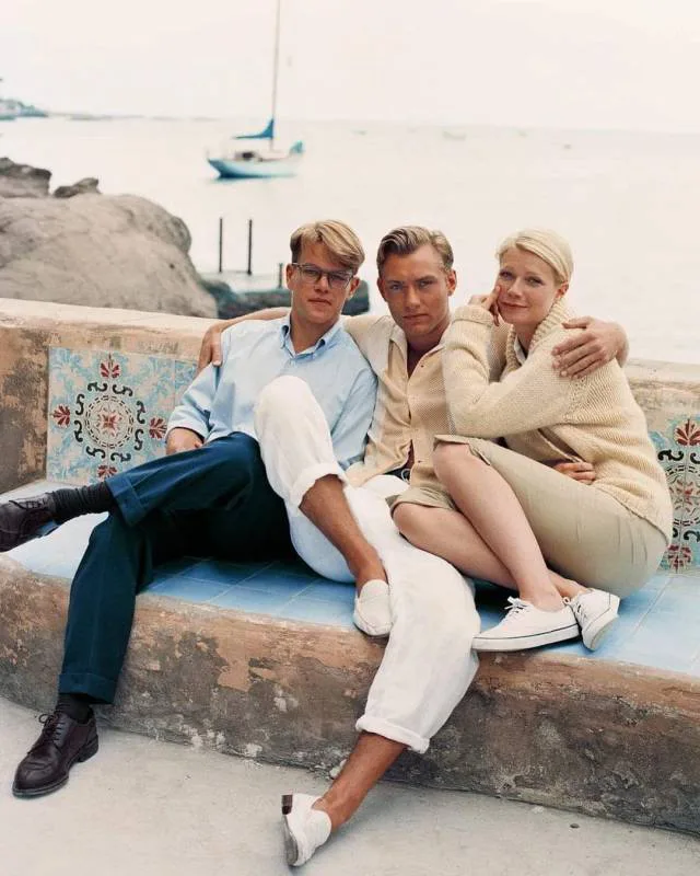 Three people sit on a seaside bench with a boat in the background; the man in the middle has his arm around the woman on his…