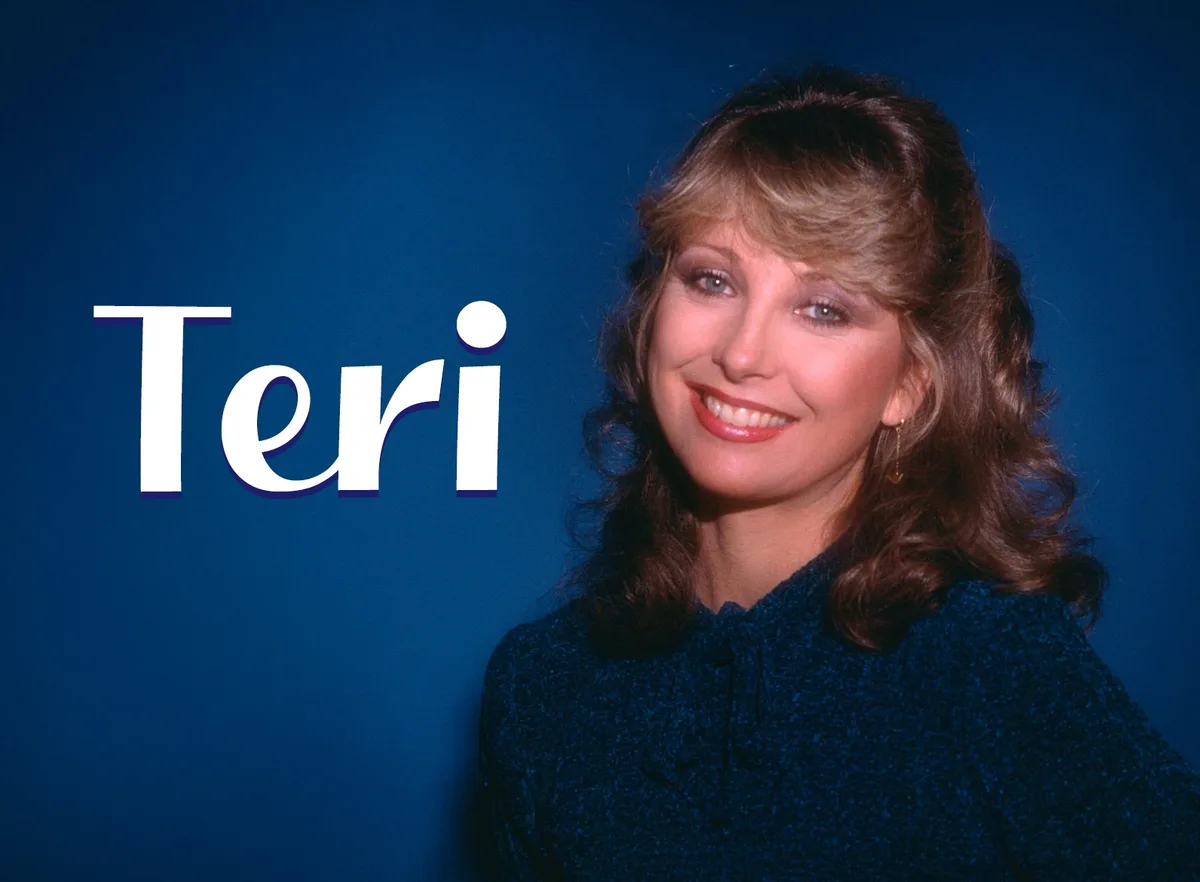 A smiling woman with voluminous, shoulder-length brown hair and bangs, wearing a dark blue textured top and dangling…