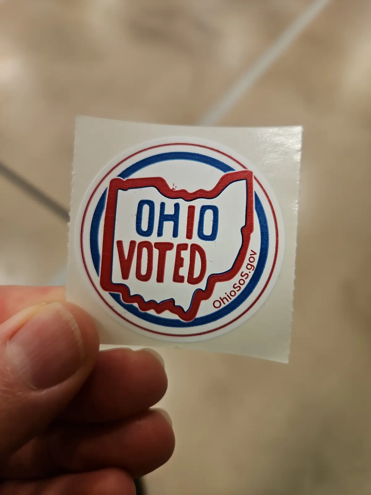 A hand holds up a circular white sticker with red and blue borders. Inside, the outline of the state of Ohio is shown in…