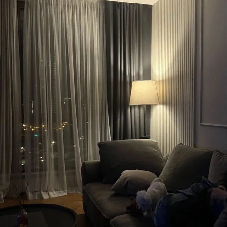 A dimly lit living room at night with a gray couch, sheer curtains revealing city lights outside, and a floor lamp casting a…
