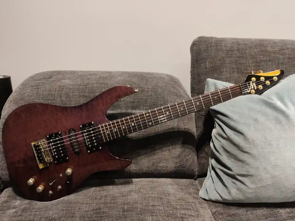 A dark reddish-brown electric guitar with a wood grain finish rests diagonally across a gray fabric couch, its headstock…