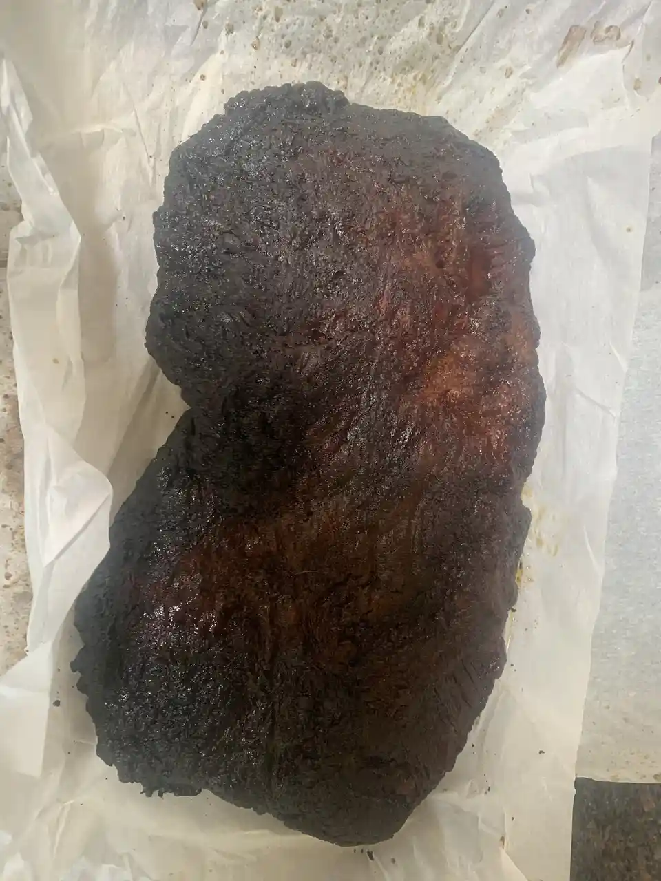 A large, heavily charred piece of meat, likely brisket or ribs, sits on white parchment paper, its surface blackened and…