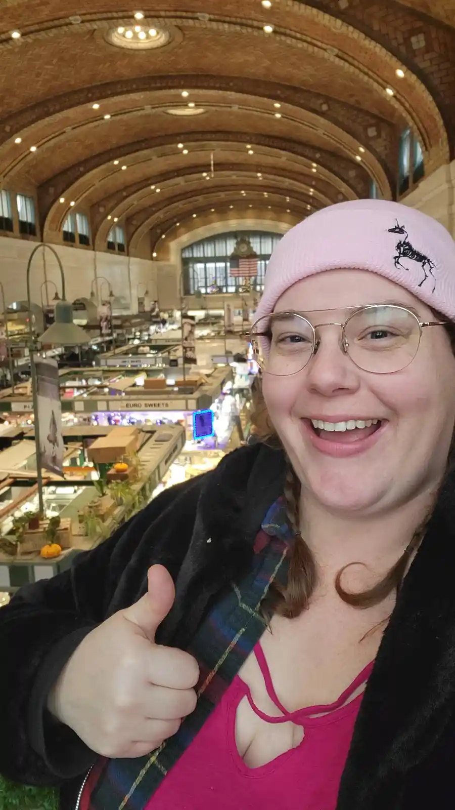 A person wearing a pink beanie with a black skeleton horse design, glasses, and a pink top gives a thumbs-up while smiling…