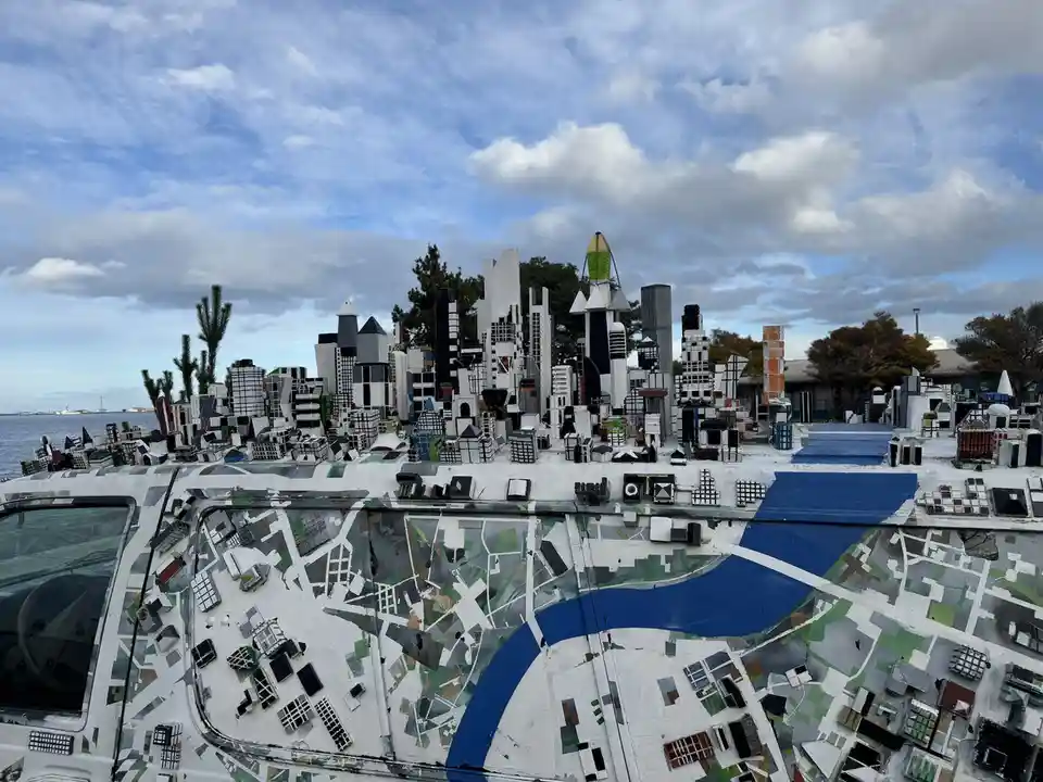A white van is heavily modified with a detailed miniature cityscape built on its roof and sides, featuring tiny buildings…