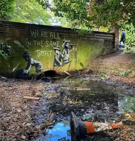 A mural painted on a mossy brick wall depicts two children in a small boat, with the text 'WE'RE ALL IN THE SAME BOAT' above…