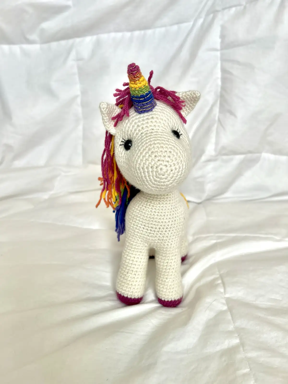 A handmade, white crocheted unicorn plush toy with a rainbow-colored horn and mane, standing on a white fabric surface. The…
