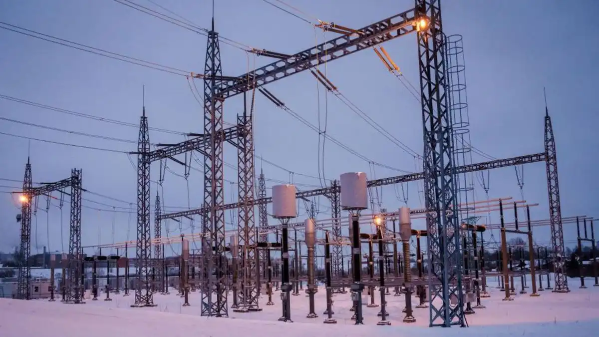 A wide shot of a snowy outdoor electrical substation at dusk, featuring tall metal lattice towers, numerous overhead power…