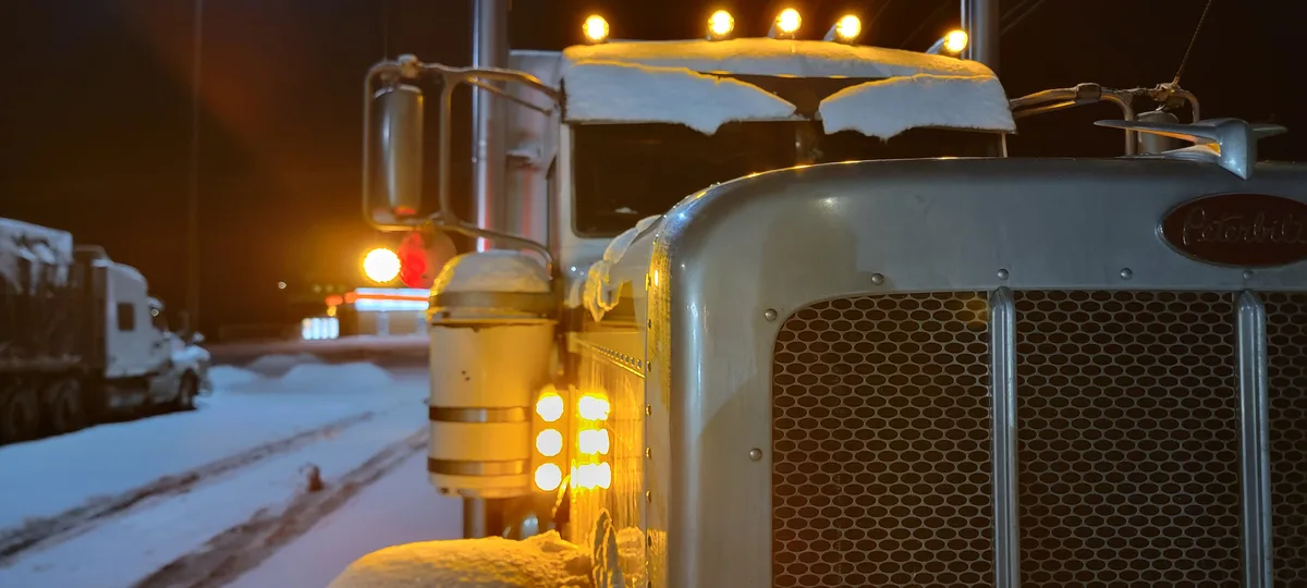 A close-up of a snow-covered Peterbilt semi-truck at night, its headlights and roof lights glowing warmly against the dark…