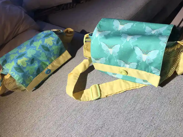 Two handmade fabric bags with yellow straps and mesh pockets sit on a gray couch. One bag has a blue background with green…