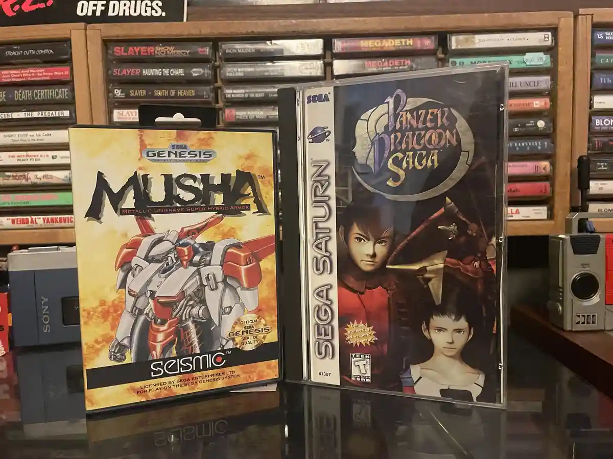 Two video game cases sit side-by-side on a glass surface: 'Musha' for the Sega Genesis, featuring a red and silver robot…
