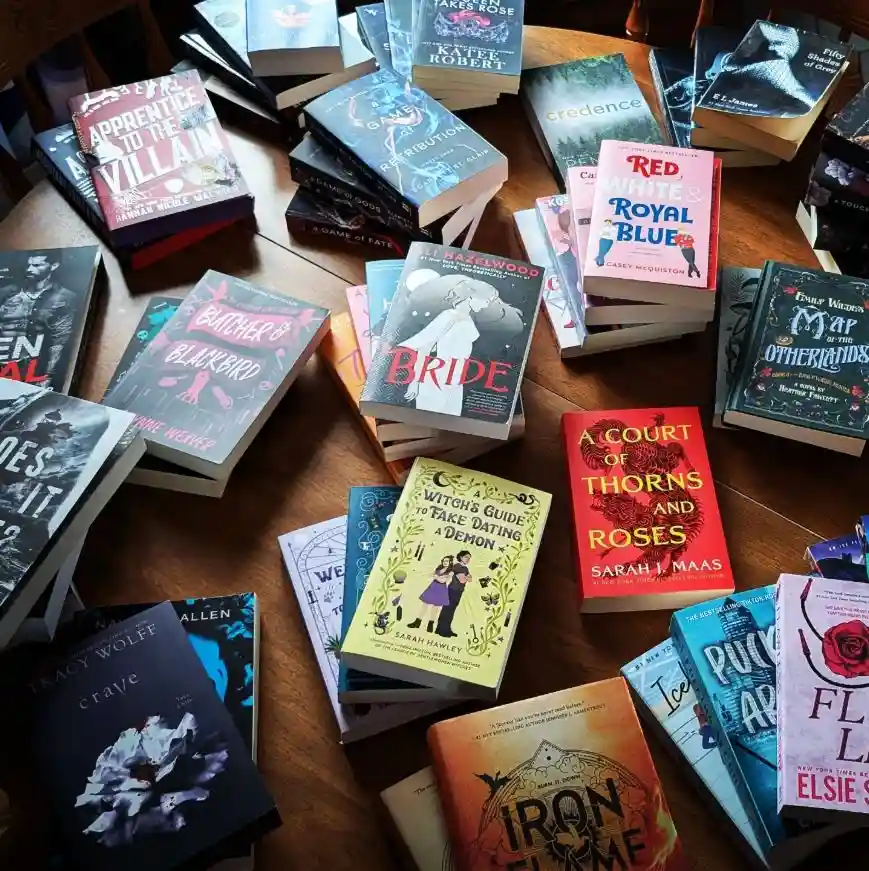 A wooden table is covered with stacks of colorful fantasy and romance novels, including titles like 'Apprentice to the…