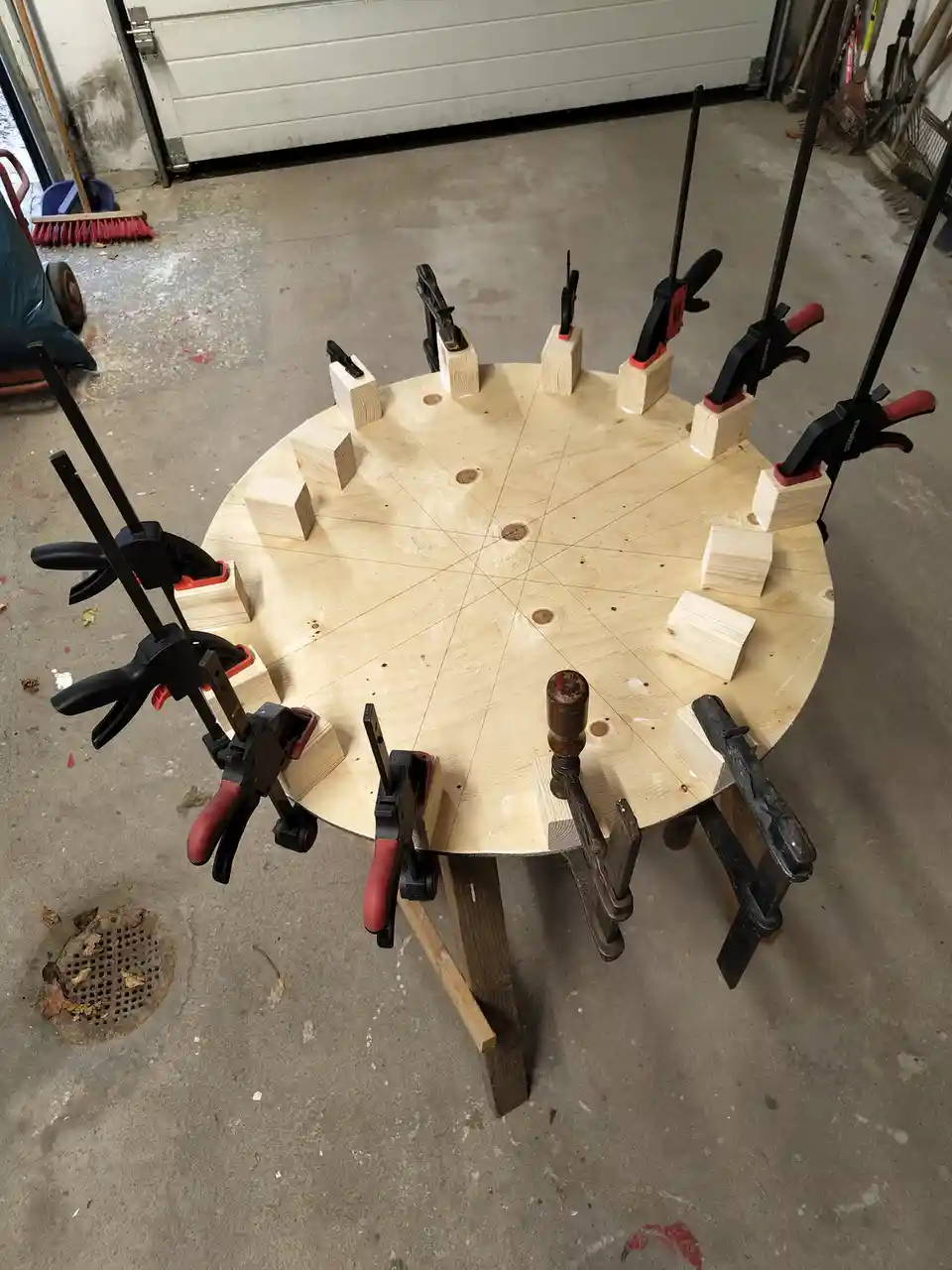 A circular wooden board is clamped with numerous clamps and wooden blocks arranged around its perimeter, set up in a garage…
