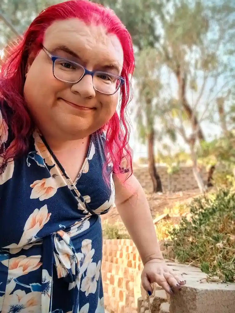 A person with bright pink hair and glasses, wearing a blue floral dress, smiles while leaning on a low wall outdoors. The…