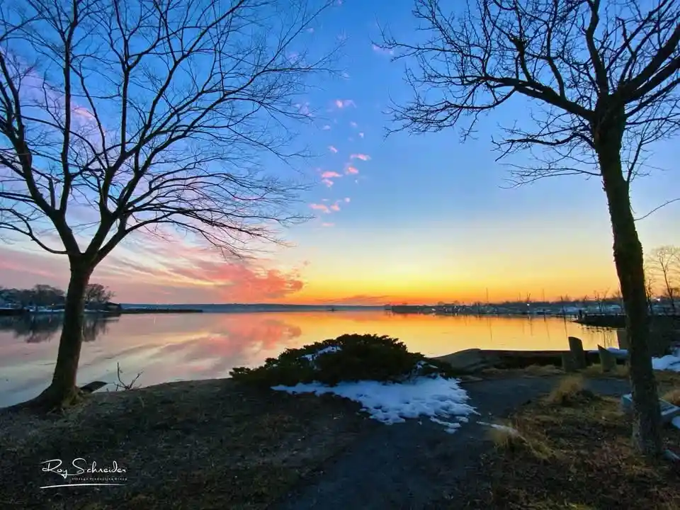 A serene winter sunset over a calm body of water, framed by two bare trees with snow patches on the ground. The sky displays…
