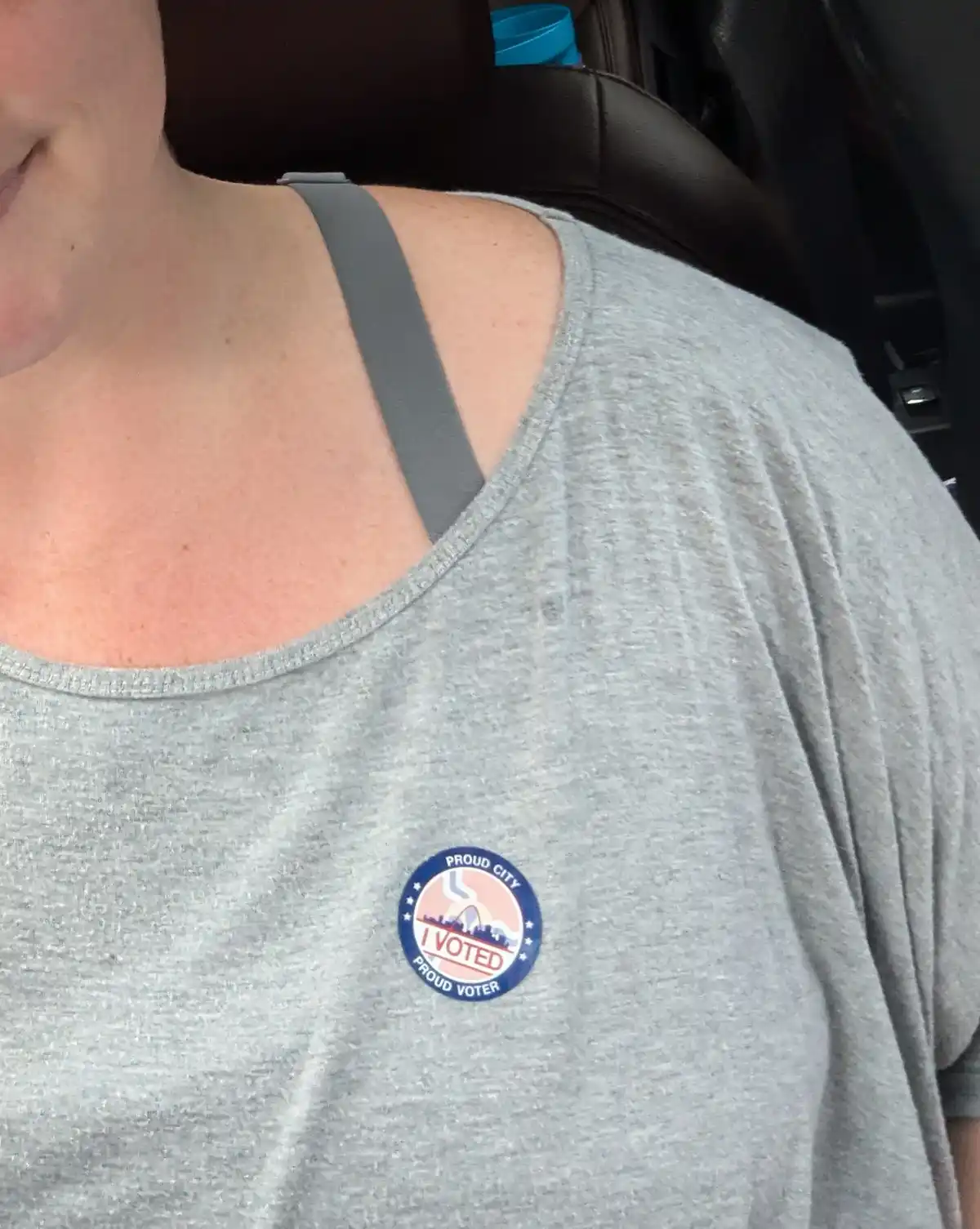 A close-up photo shows a person wearing a gray t-shirt with a circular 'I VOTED' sticker pinned to the chest. The sticker…