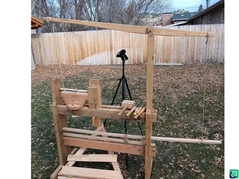 A homemade wooden catapult sits in a backyard, constructed from planks and ropes, with a camera on a tripod positioned to…