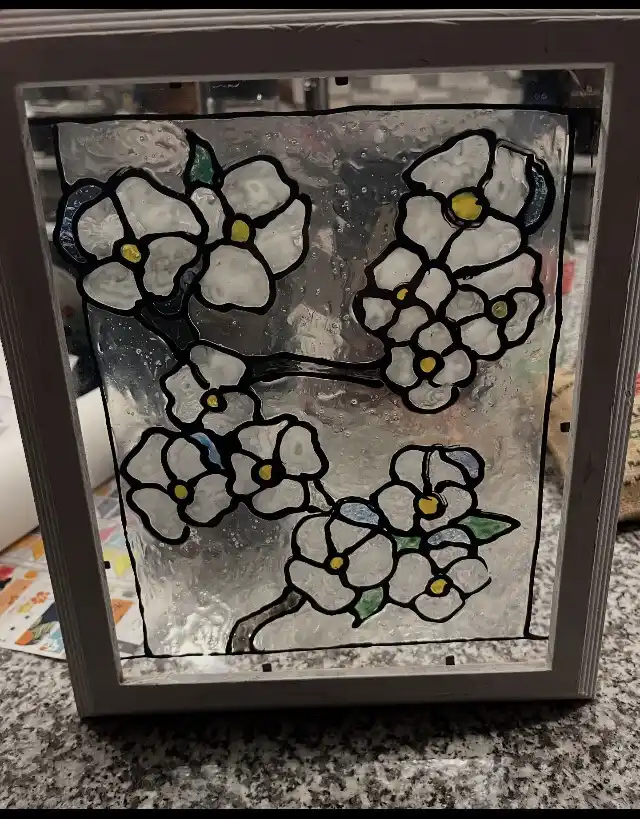 A handmade stained glass panel depicting white flowers with yellow centers and green leaves, set in a simple gray frame…