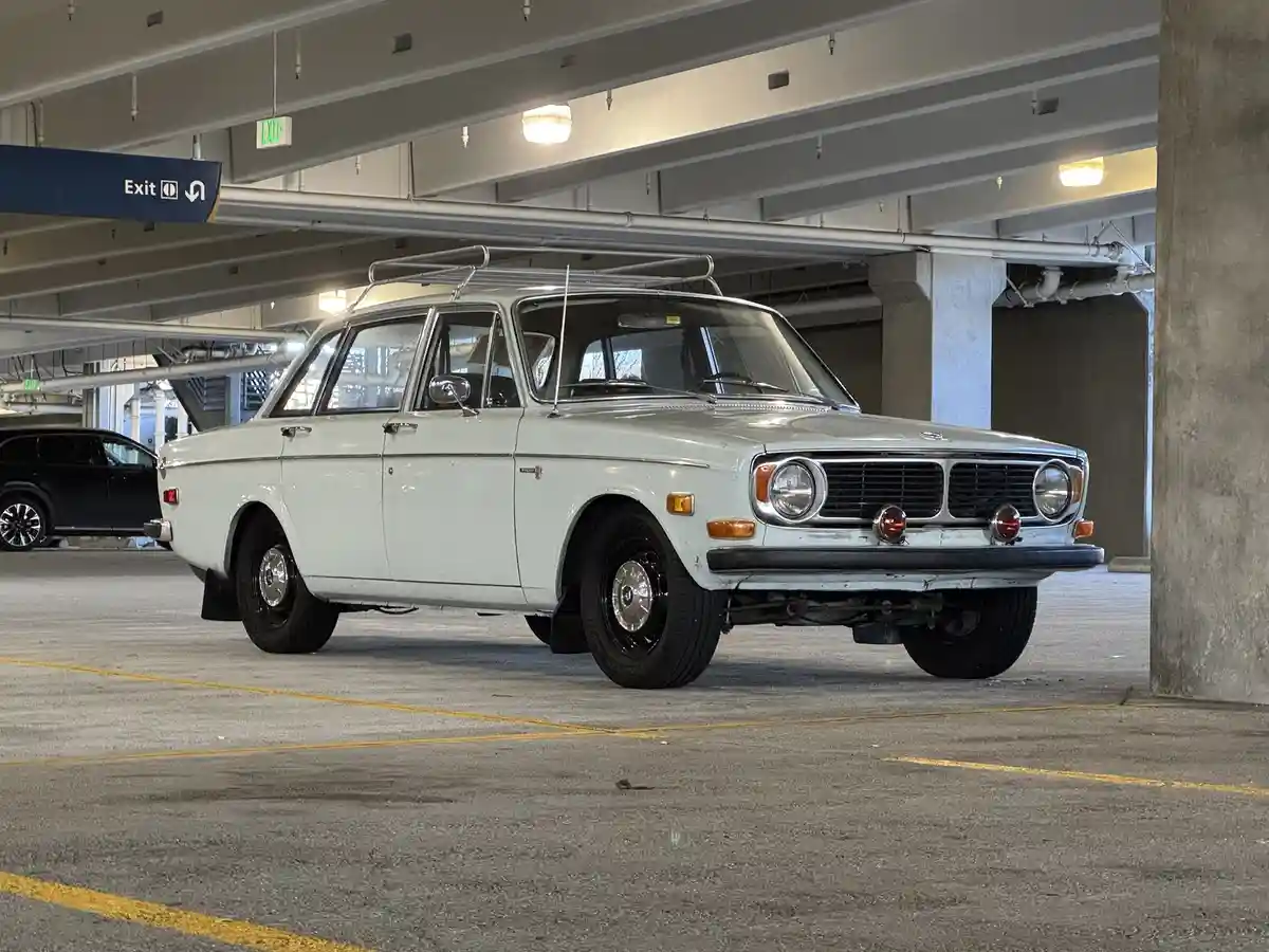 A light-colored classic Volvo station wagon with a roof rack is parked in a modern concrete parking garage, with an 'Exit'…