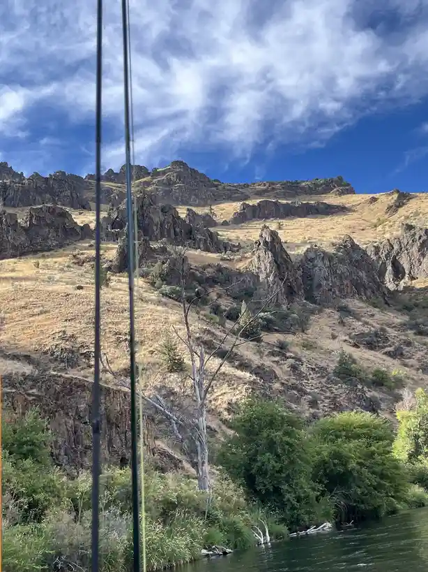 A scenic photo of a river canyon with rocky hills, dry grass, and green trees under a partly cloudy blue sky, viewed through…