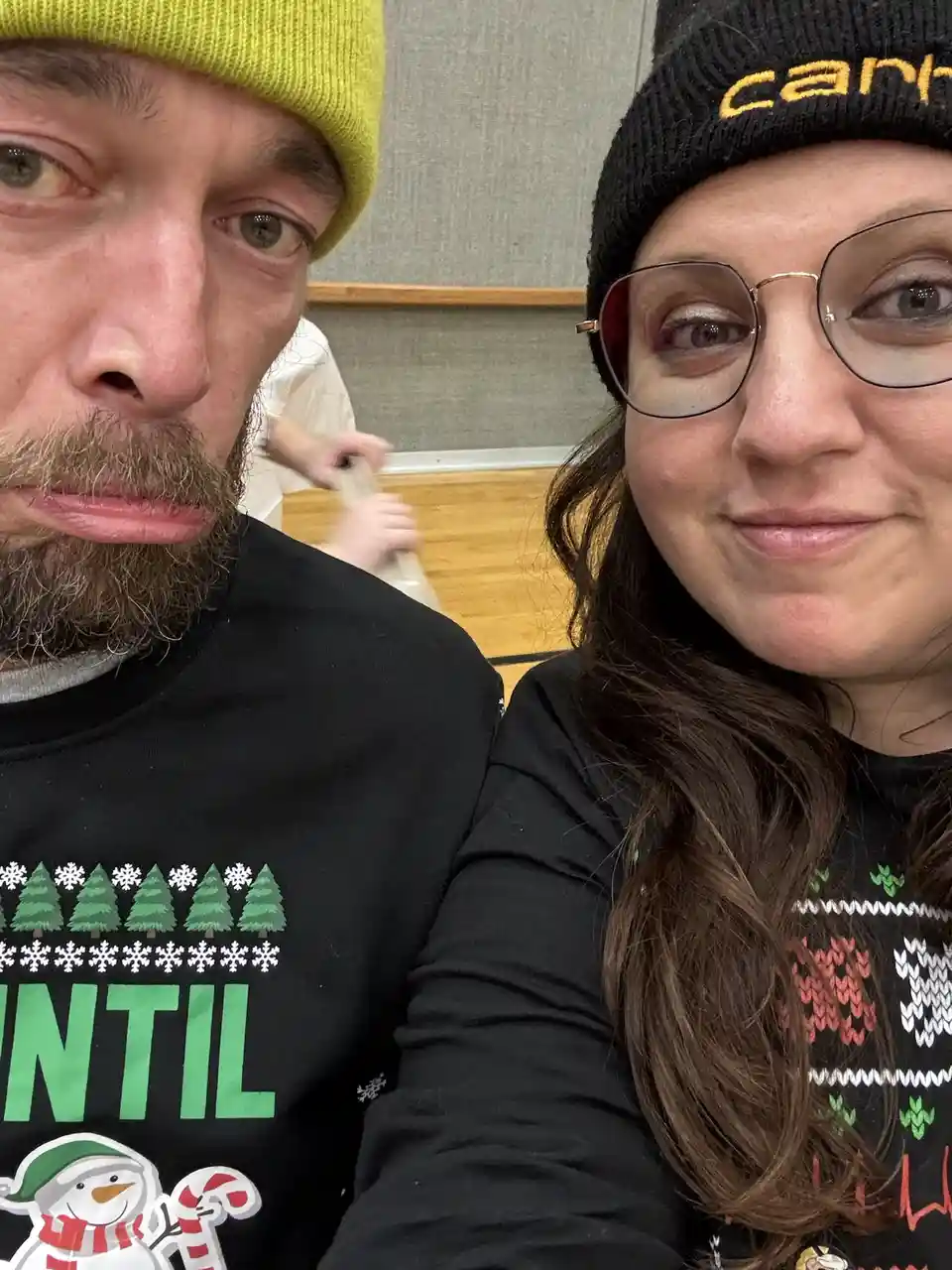 A selfie of a man and woman in festive christmas sweaters, the man on the left wearing a lime green beanie and making a…