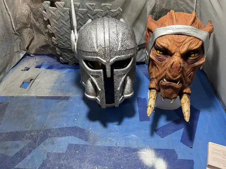 Two custom-made fantasy helmets sit side-by-side on a blue painter's tarp. The left helmet is silver with intricate swirling…