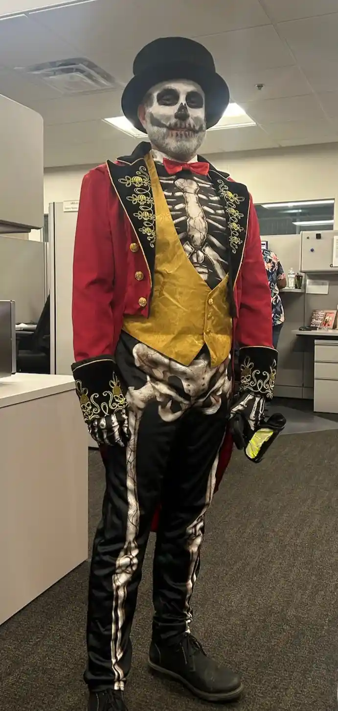 A person in an office wearing a full skeleton costume with a red jacket, gold vest, black pants, and top hat, holding a…