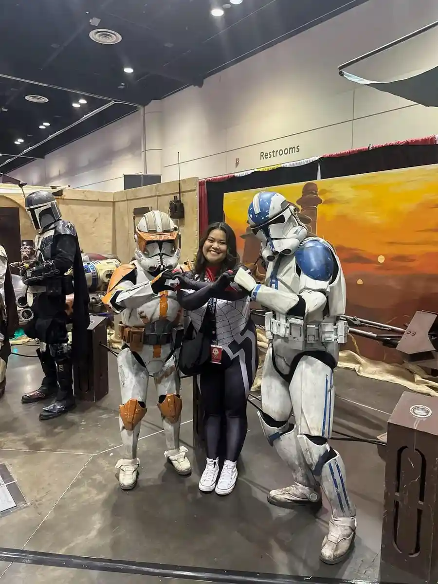A woman in a cosplay outfit stands between two Star Wars clone troopers at a convention, both troopers are holding their…