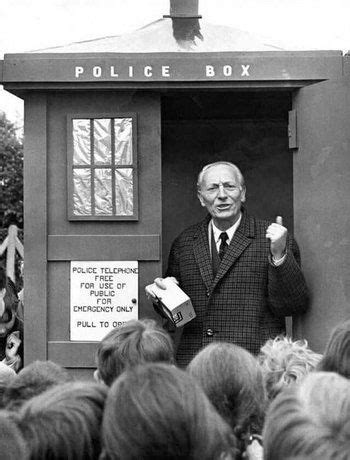 A black-and-white photo shows an older man in a suit standing inside a classic British police box, holding a small box and…
