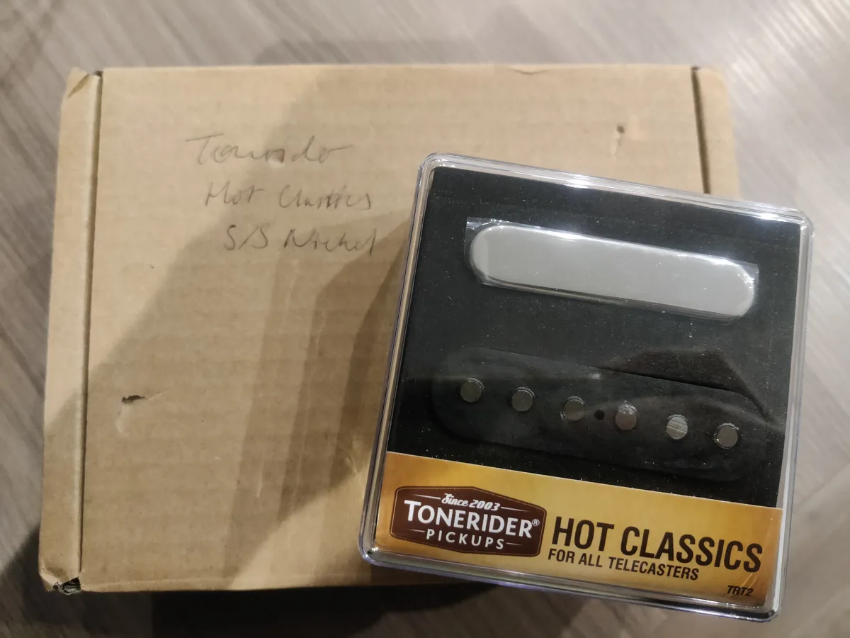 A close-up photo shows a Tonerider Hot Classics guitar pickup in its clear plastic packaging, sitting next to a plain brown…