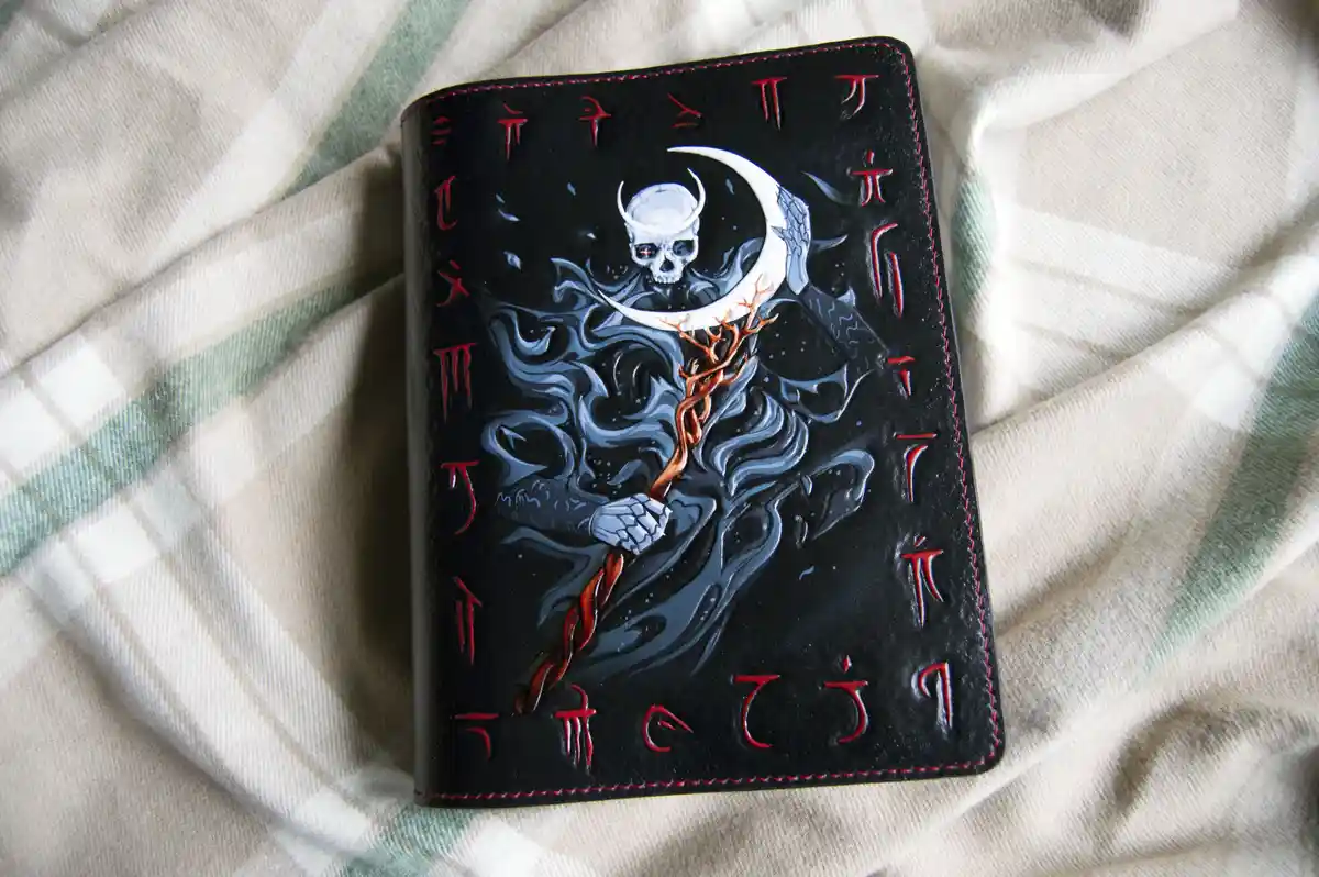 A black leather-bound notebook with red stitching lies on a plaid fabric. The cover features a detailed illustration of a…